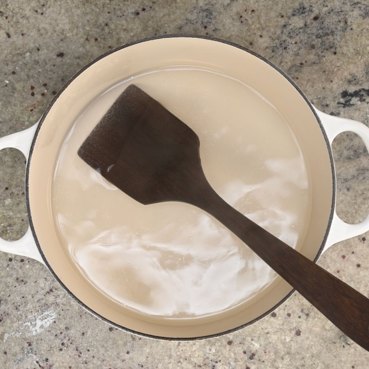 Clear pickling brine solution being stirred with a wooden spatula in a white pot.