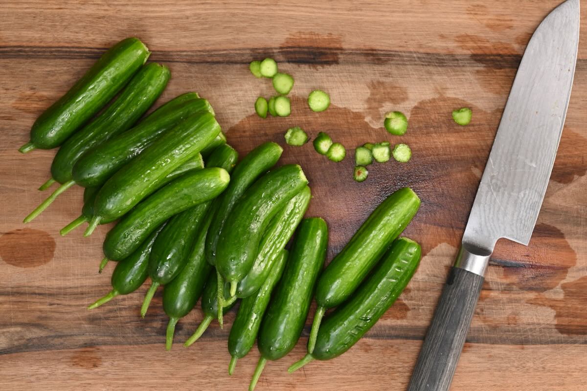 Fresh small green cucumbers on a wooden cutting board with trimmed stem ends beside a chef's knife.