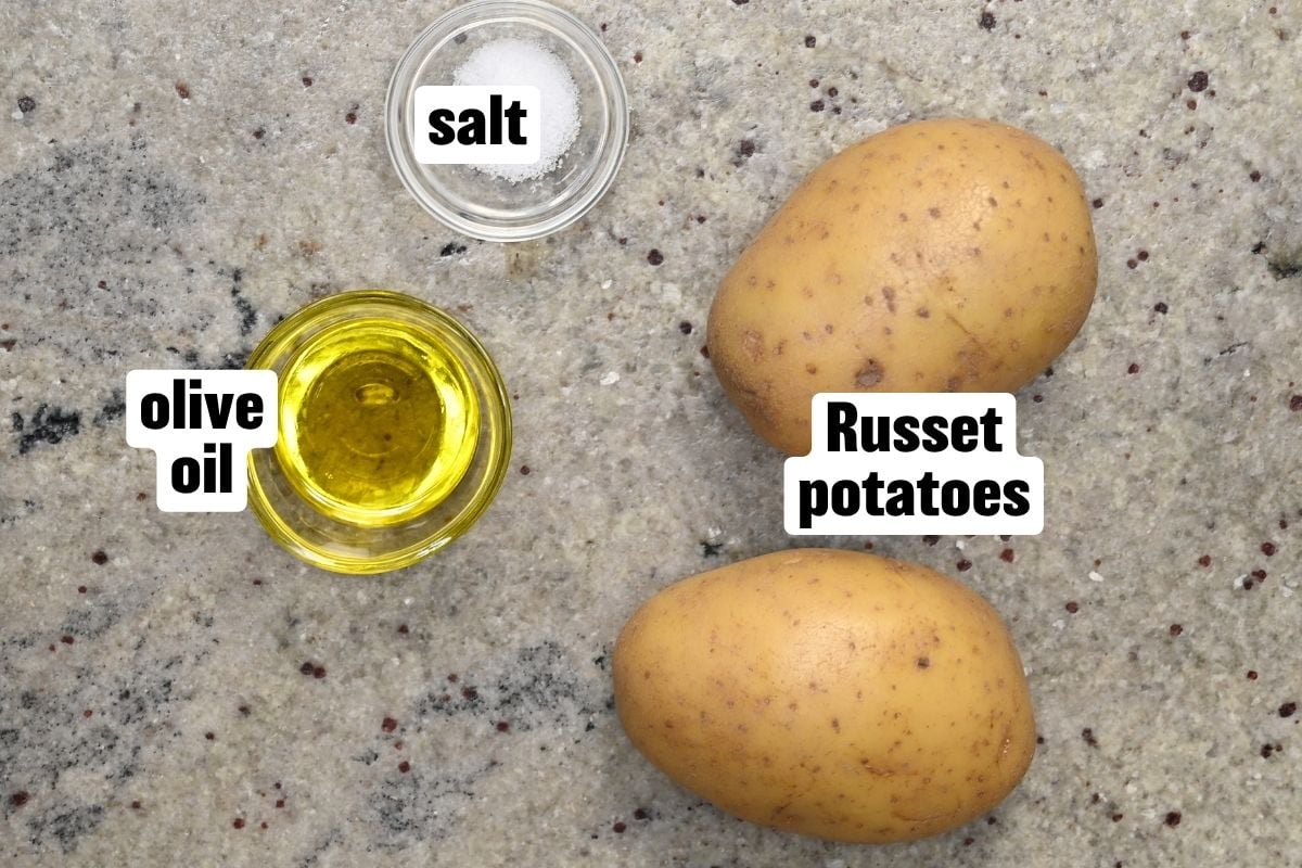 Top-down view of ingredients for shoestring fries arranged on a countertop, including two whole Russet potatoes, a small glass dish of olive oil, and a small glass dish of salt.