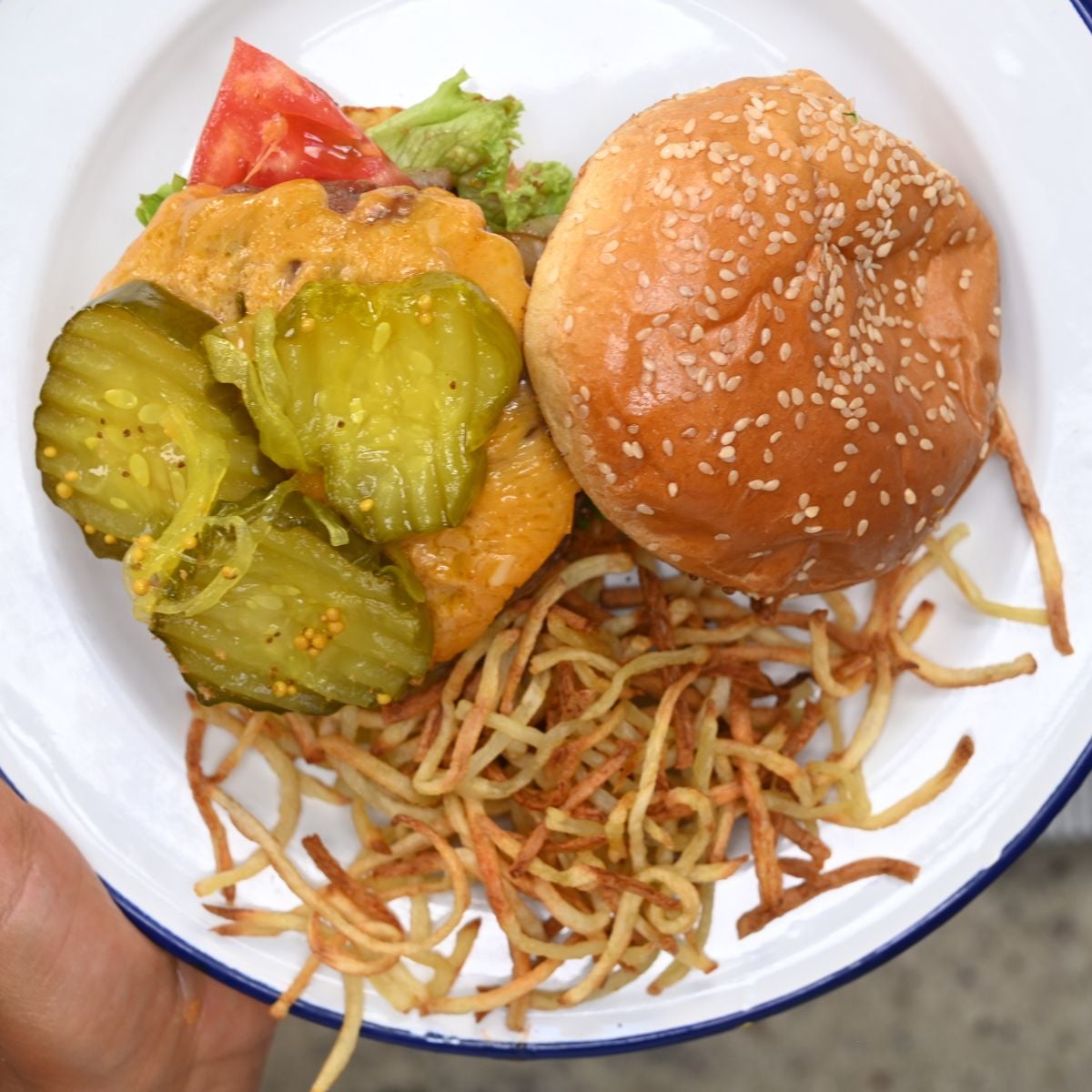 Top-down view of an open-faced cheeseburger topped with pickles, served alongside a pile of crisp shoestring fries on a white enamel plate.