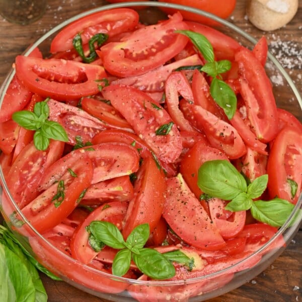 Close-up overhead view of seasoned tomato salad made with red tomato wedges, sea salt, black pepper, and fresh basil sprigs served in a shallow glass bowl.