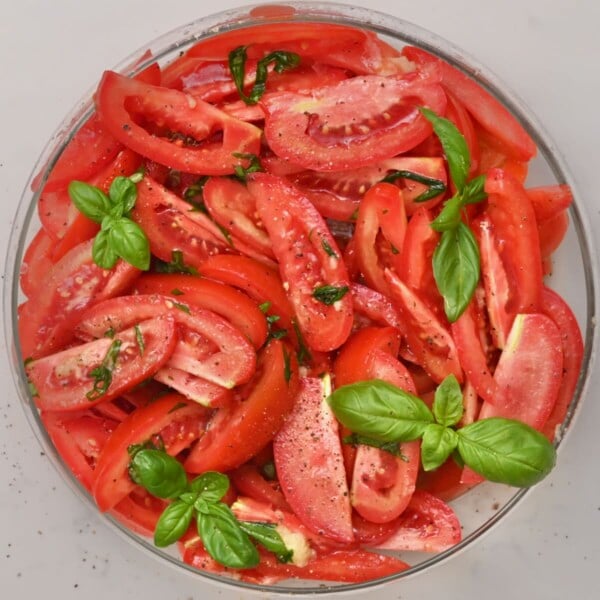 Overhead shot of freshly tossed tomato salad in a glass bowl coated in olive oil dressing with fresh basil leaves mixed throughout.
