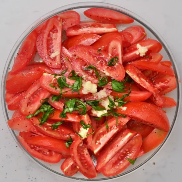 Overhead view of sliced tomatoes in a glass bowl topped with minced garlic, chopped fresh basil, olive oil, and black pepper before tossing.