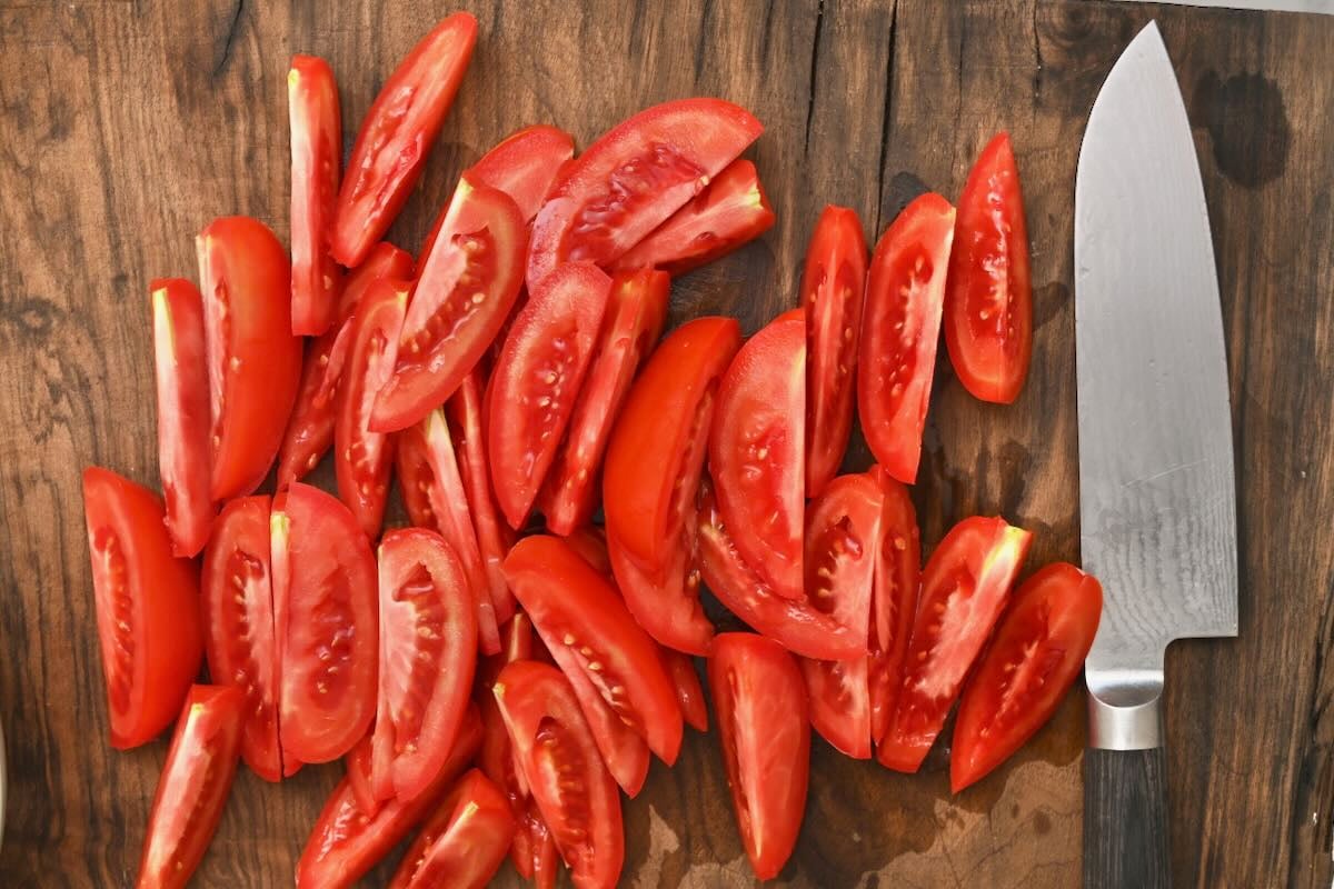 Sliced ripe Roma tomato wedges piled on a dark wooden cutting board next to a sharp chef's knife.