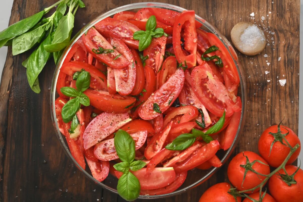 Top-down view of a completed tomato salad in a clear bowl on a wooden board surrounded by fresh basil leaves, vine tomatoes, and sea salt.