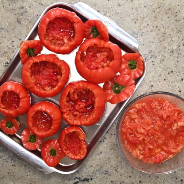 Hollowed-out red tomatoes and their trimmed tops arranged on a metal baking tray next to a bowl of tomato pulp.