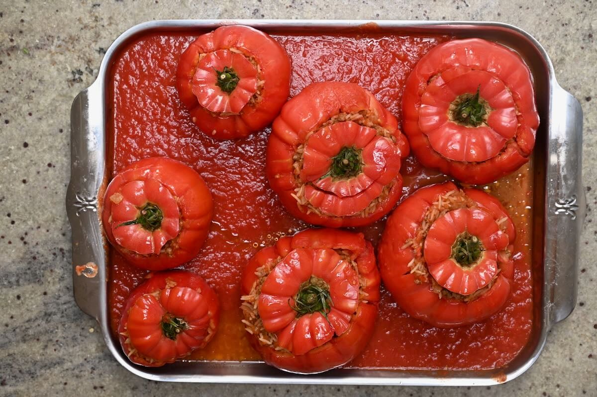 Rice-stuffed tomatoes topped with tomato caps and surrounded by tomato sauce on a tray before baking.