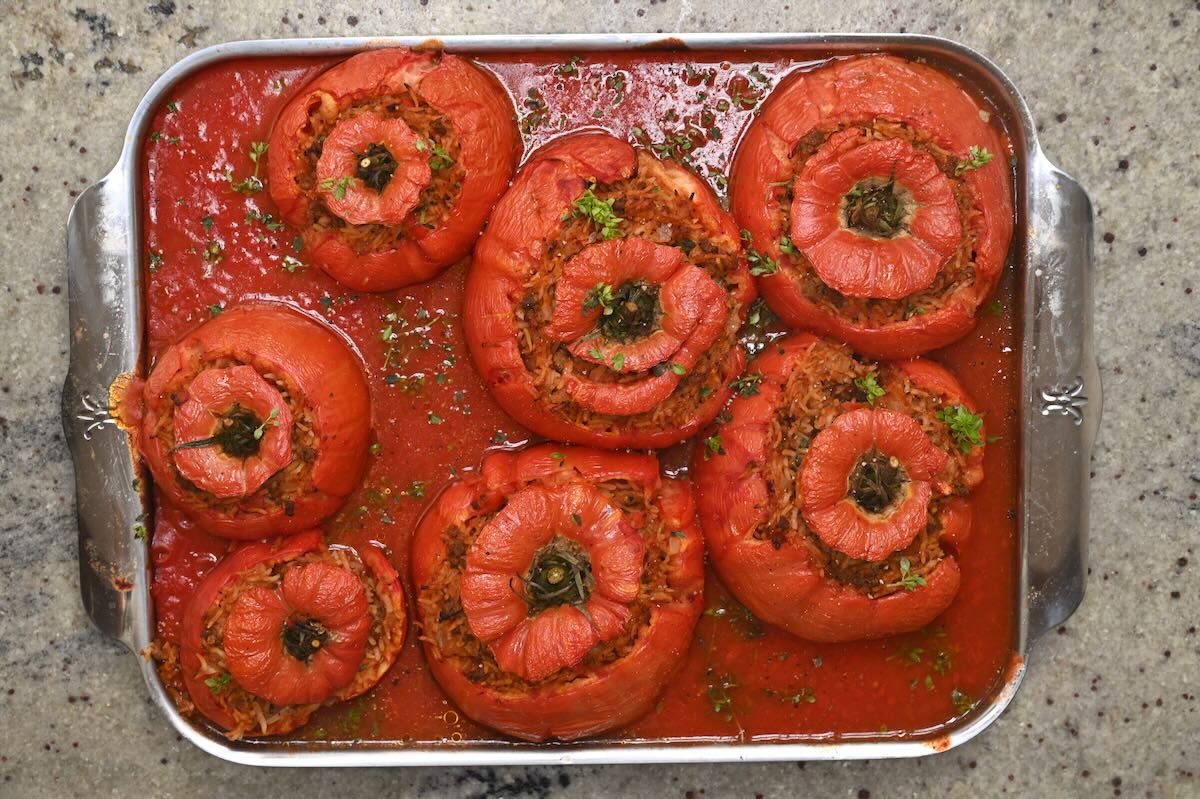 Top-down view of baked stuffed tomatoes topped with fresh herbs in tomato sauce on a metal baking dish.