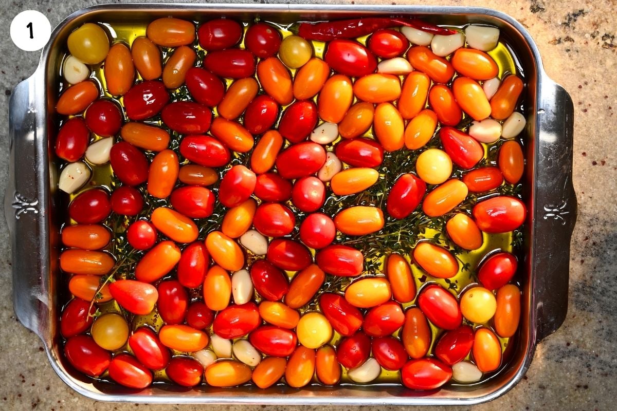 Cherry tomatoes, garlic cloves, thyme sprigs, and red chili submerged in olive oil in a stainless steel baking dish before roasting.