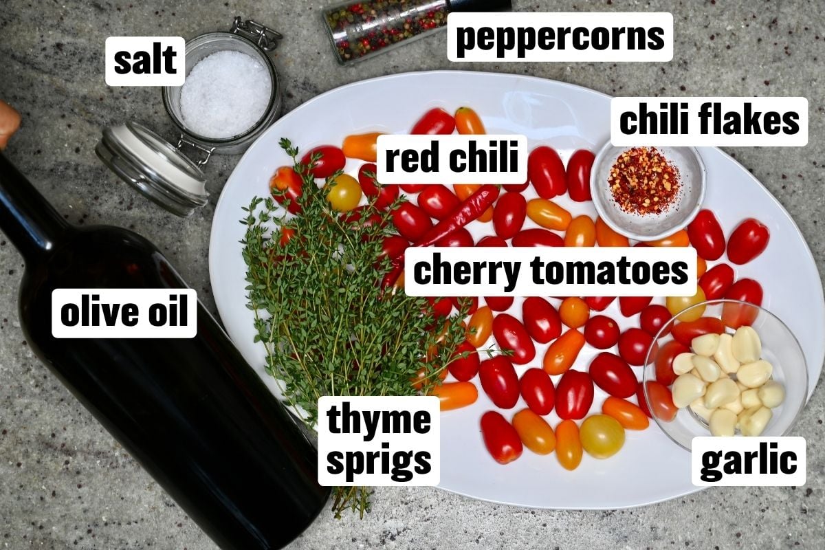 A white oval platter surrounded by ingredients for tomato confit, including cherry tomatoes, thyme sprigs, garlic, red chili, chili flakes, olive oil, salt, and peppercorns.
