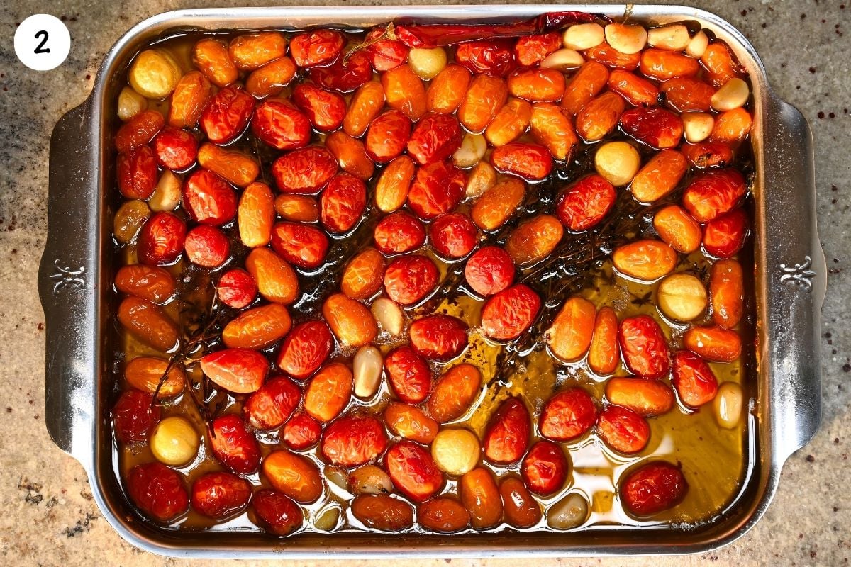 Slow-roasted cherry tomatoes, softened garlic, thyme sprigs, and red chili submerged in warm olive oil in a metal baking dish.