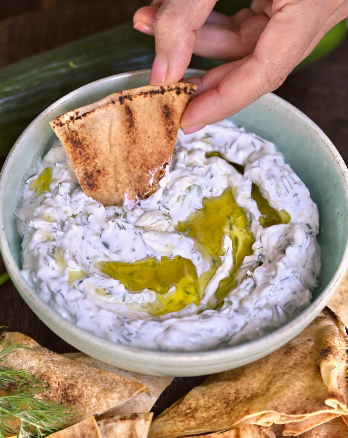 Hand dipping a piece of grilled pita bread into a bowl of creamy authentic tzatziki sauce swirled with olive oil.