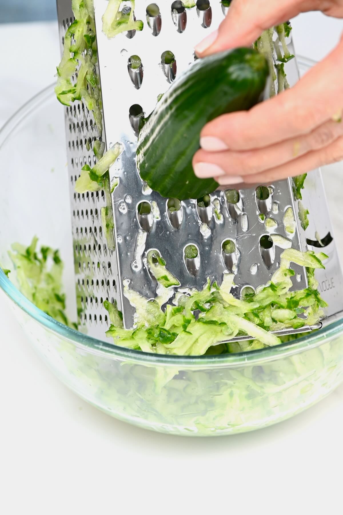 Hand grating a fresh English cucumber into a glass bowl using a stainless steel box grater.