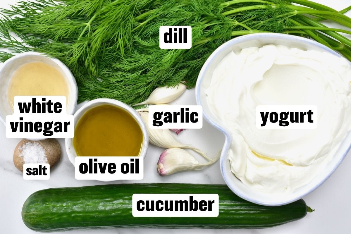 Overhead view of authentic tzatziki ingredients with text labels including cucumber, Greek yogurt, fresh dill, garlic, olive oil, white vinegar, and salt.