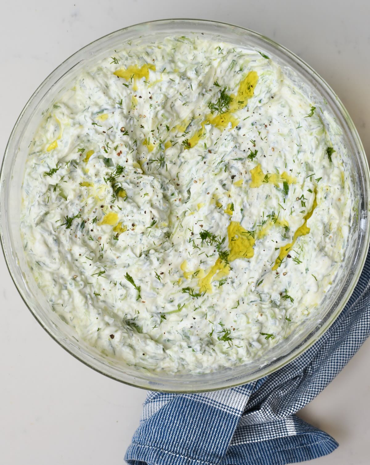 Overhead view of authentic homemade tzatziki sauce in a clear glass bowl resting next to a blue patterned kitchen towel.