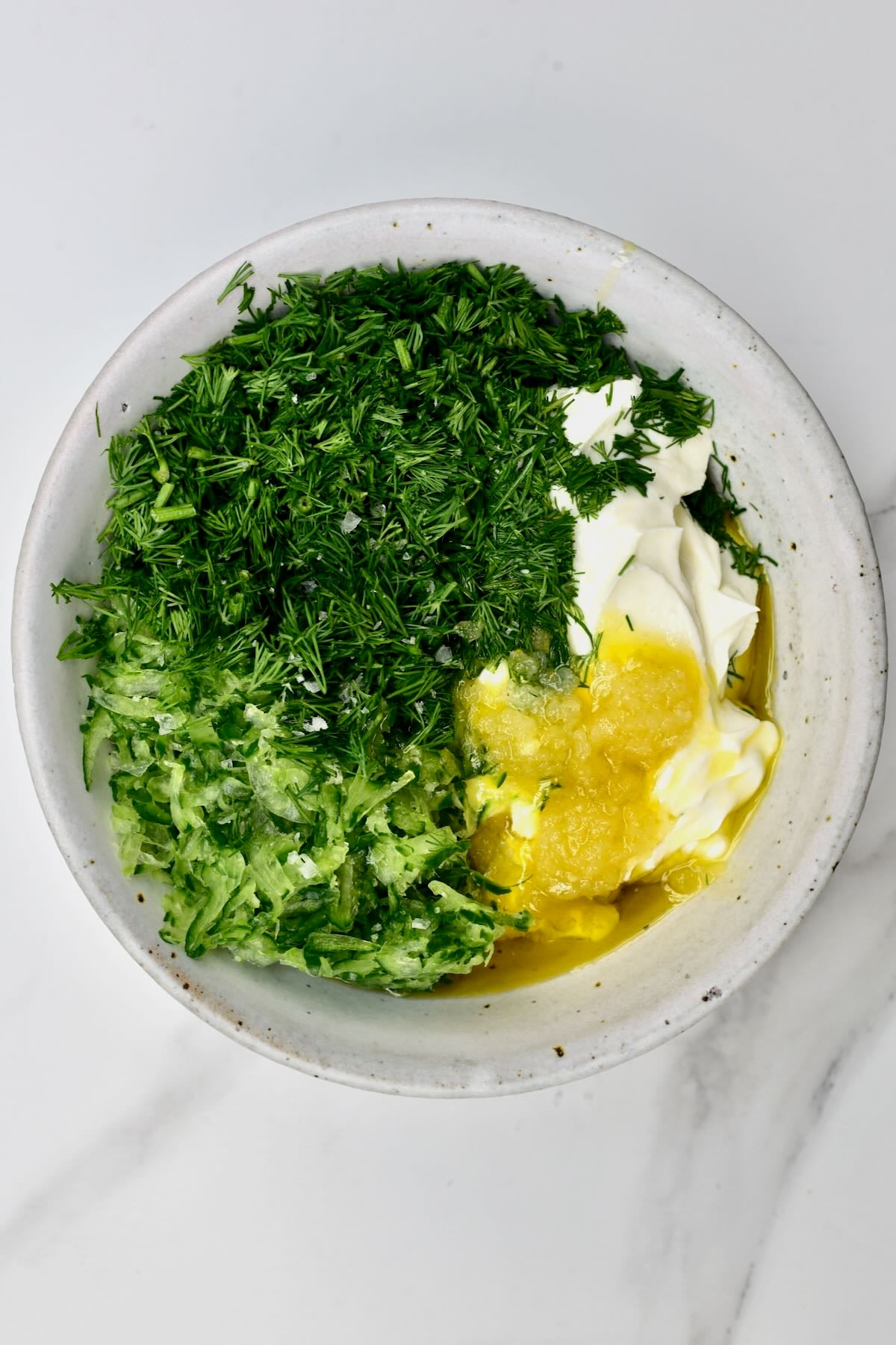 Mixing bowl containing Greek yogurt, drained grated cucumber, finely chopped fresh dill, minced garlic, olive oil, and vinegar before stirring.