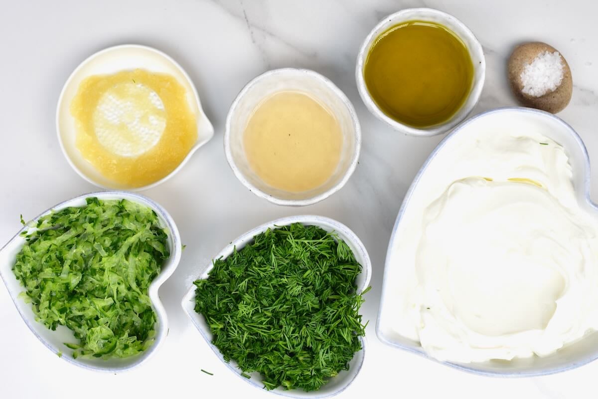 Small ceramic bowls containing prepped ingredients for tzatziki including squeezed grated cucumber, chopped fresh dill, minced garlic, olive oil, vinegar, salt, and Greek yogurt.