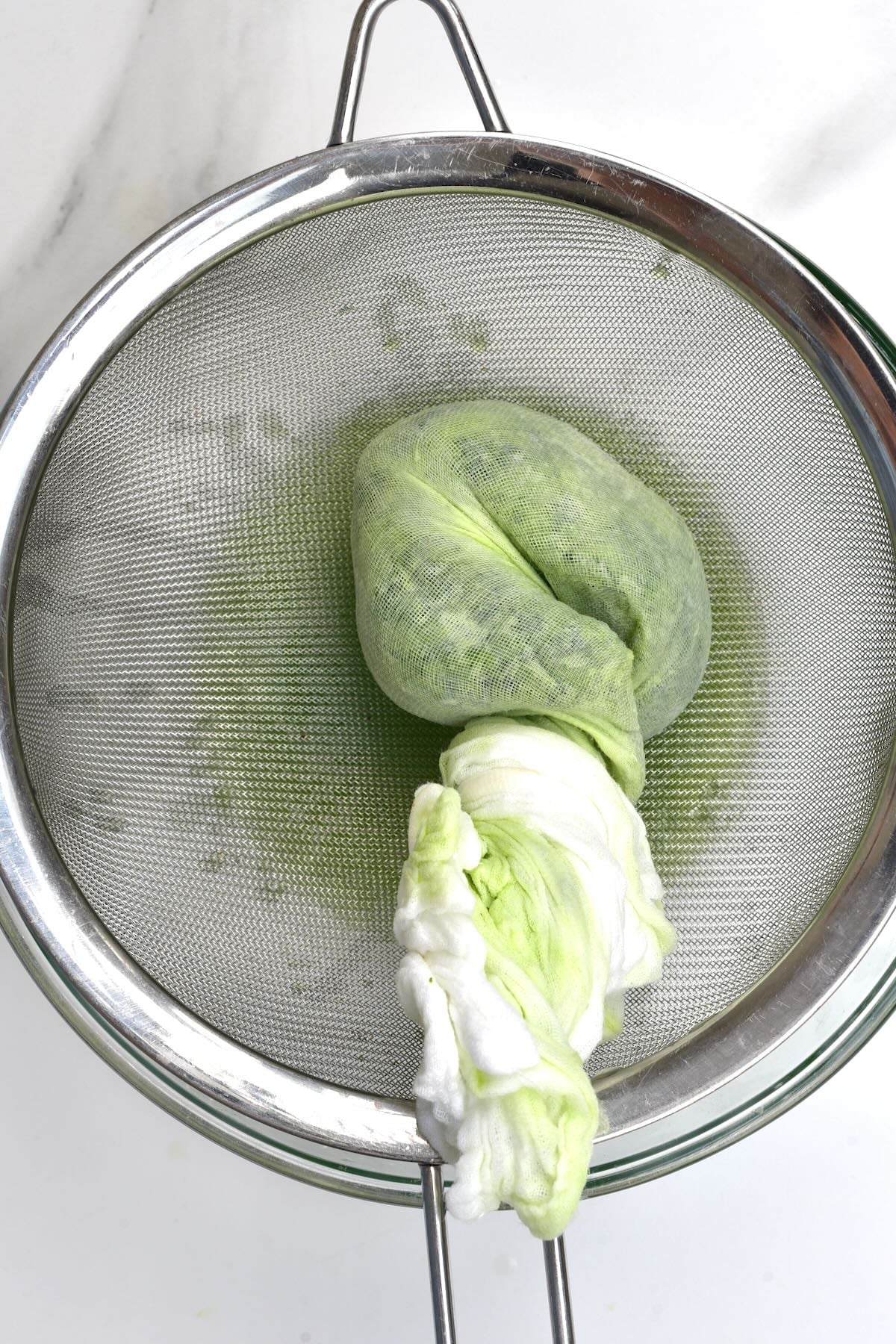 Squeezing excess liquid from salted grated cucumber using a cheesecloth over a fine-mesh strainer.