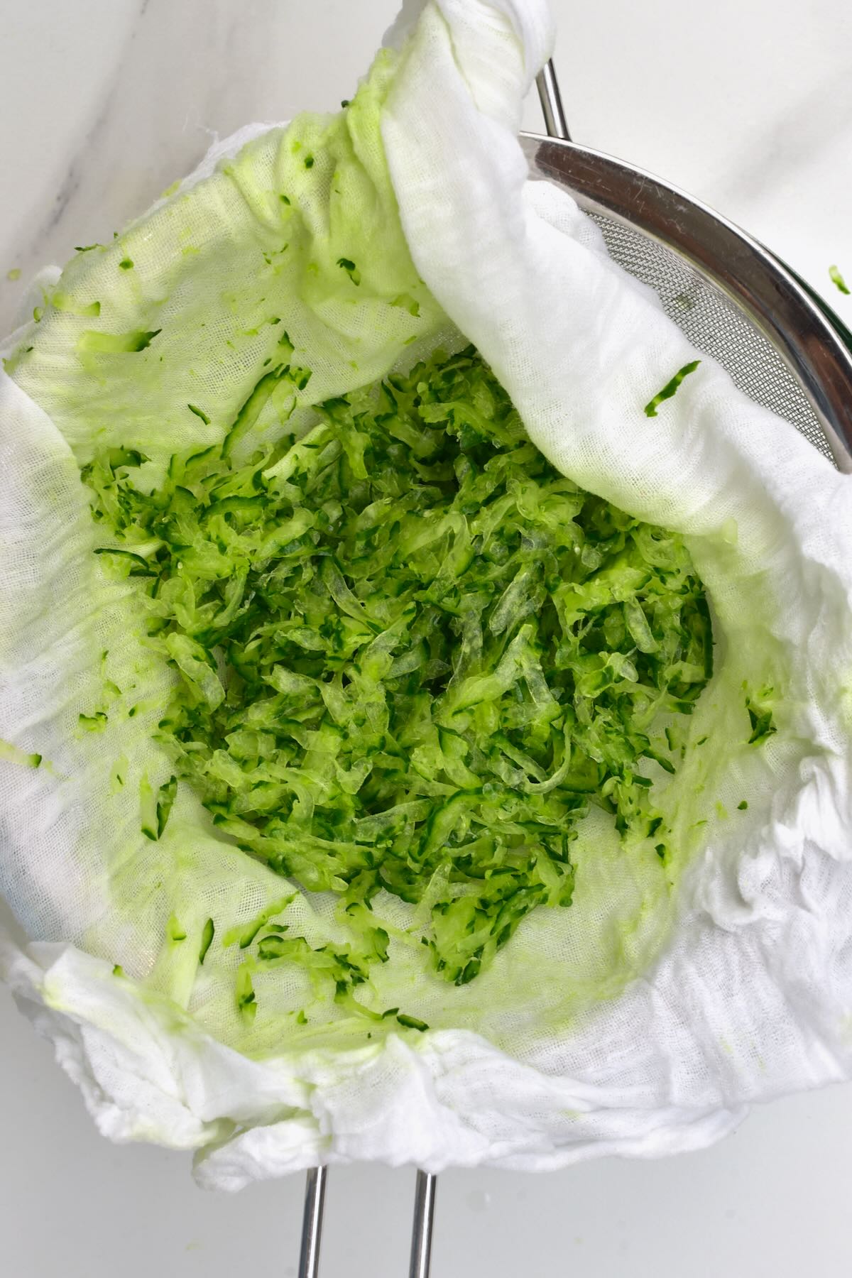 Well-drained grated cucumber resting inside a white cheesecloth set over a sieve.