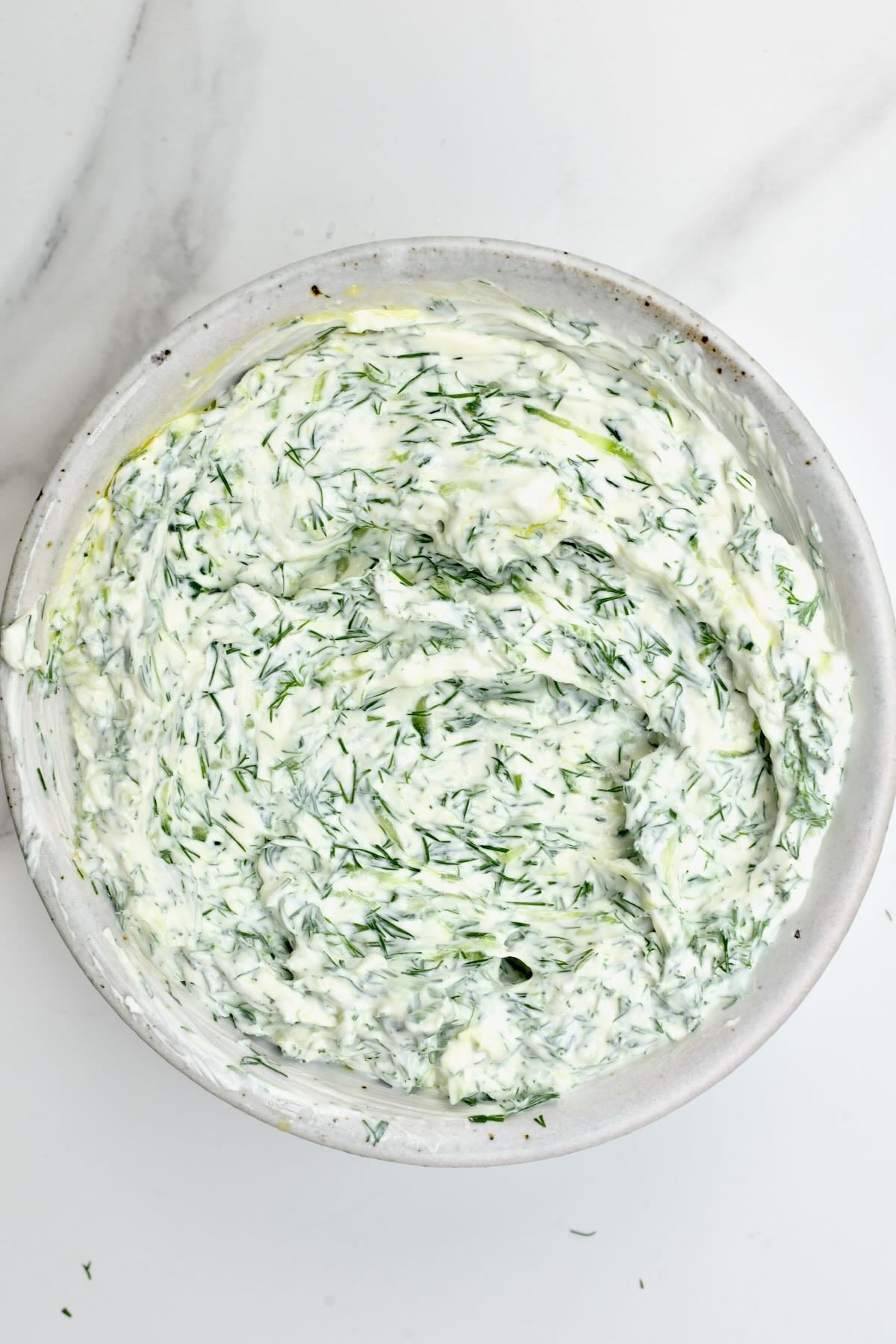 White ceramic bowl filled with freshly mixed creamy cucumber tzatziki sauce with chopped dill throughout.