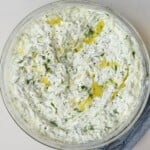 Square overhead view of a large glass bowl filled with creamy Greek tzatziki sauce garnished with extra virgin olive oil and chopped dill.