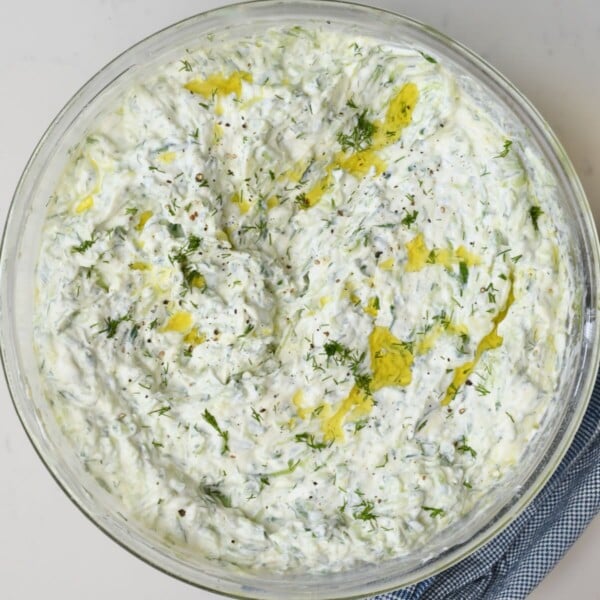Square overhead view of a large glass bowl filled with creamy Greek tzatziki sauce garnished with extra virgin olive oil and chopped dill.