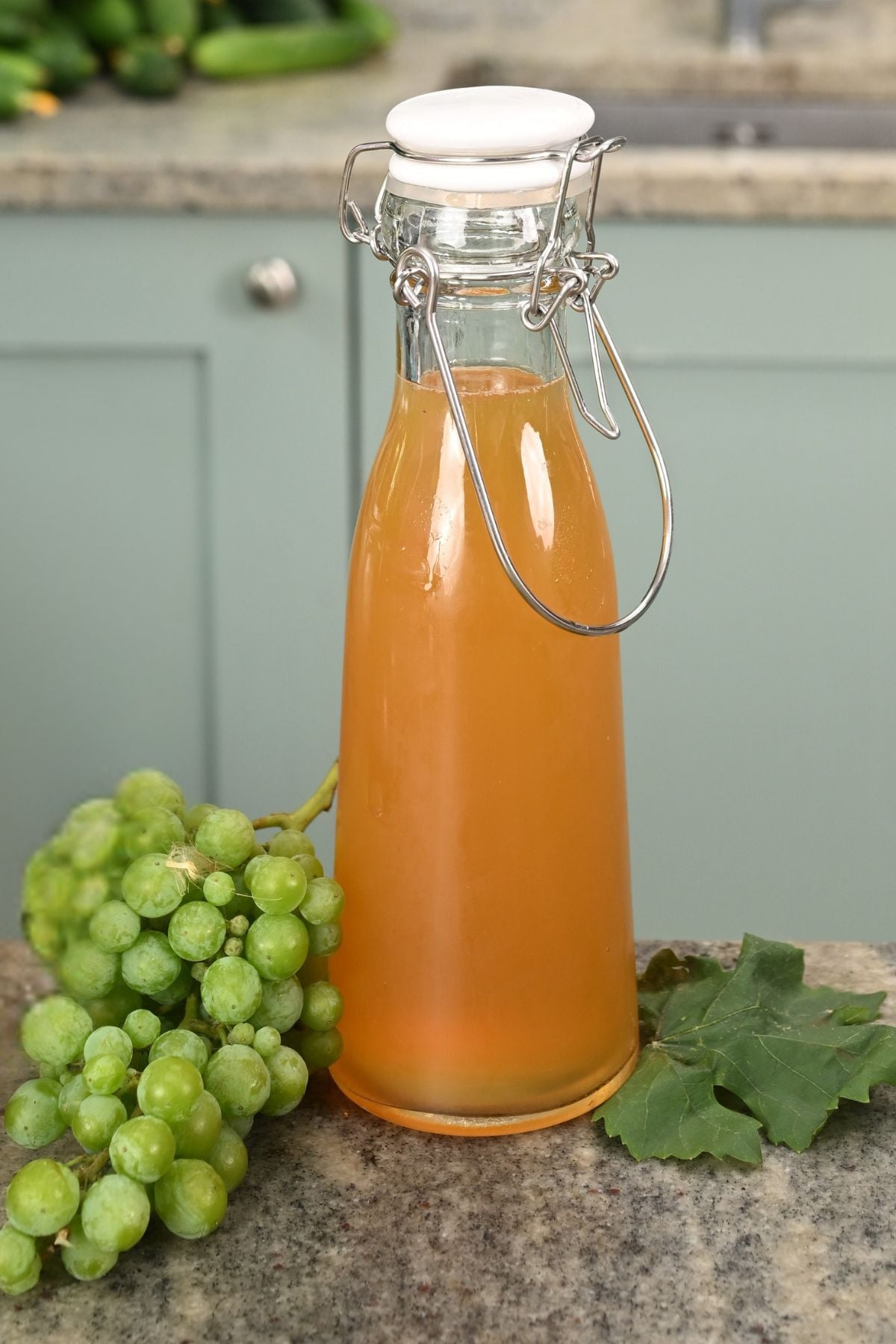 A glass swing-top bottle filled with golden verjuice, sitting on a granite countertop next to a cluster of fresh green grapes and a grape leaf.