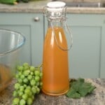A clear swing-top glass bottle filled with golden-amber verjuice, sitting on a granite countertop next to a cluster of green grapes, a grape leaf, and a mixing bowl.