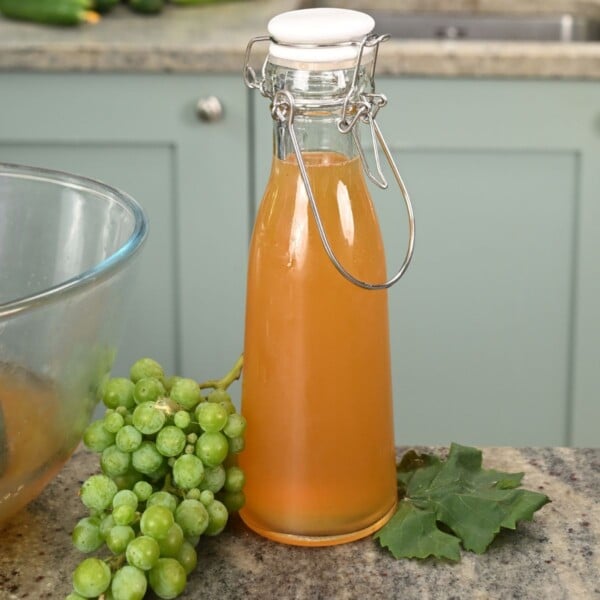A clear swing-top glass bottle filled with golden-amber verjuice, sitting on a granite countertop next to a cluster of green grapes, a grape leaf, and a mixing bowl.