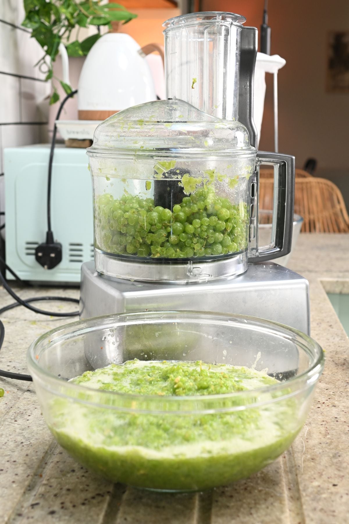 A food processor filled with fresh green grapes sitting on a countertop, with a large glass bowl of blended green grape pulp in front of it.