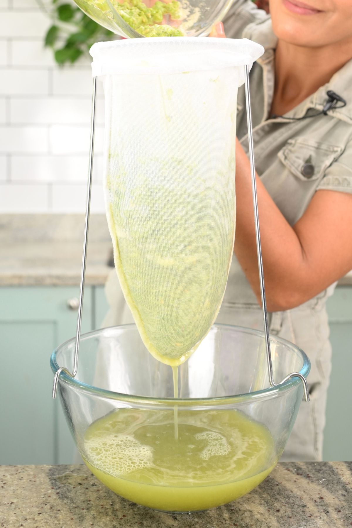 Samira pouring blended green grape mixture into a hanging mesh jelly bag to strain the fresh verjuice into a clear glass bowl below.