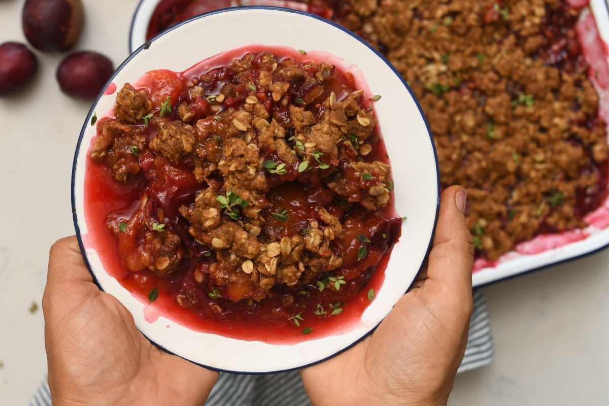 Hands holding a plate of warm plum crisp with golden oat topping and fresh thyme leaves over a baking dish on a light surface.