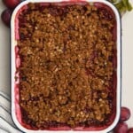 Top-down view of a baking dish filled with homemade plum crisp and a crunchy golden oat topping, framed by fresh plums on a marble surface.