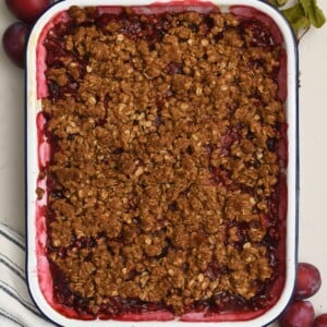 Top-down view of a baking dish filled with homemade plum crisp and a crunchy golden oat topping, framed by fresh plums on a marble surface.