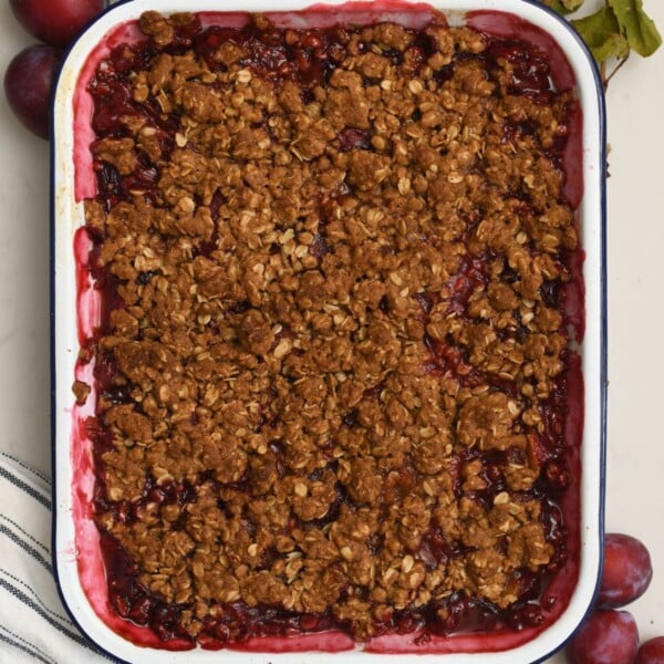 Top-down view of a baking dish filled with homemade plum crisp and a crunchy golden oat topping, framed by fresh plums on a marble surface.