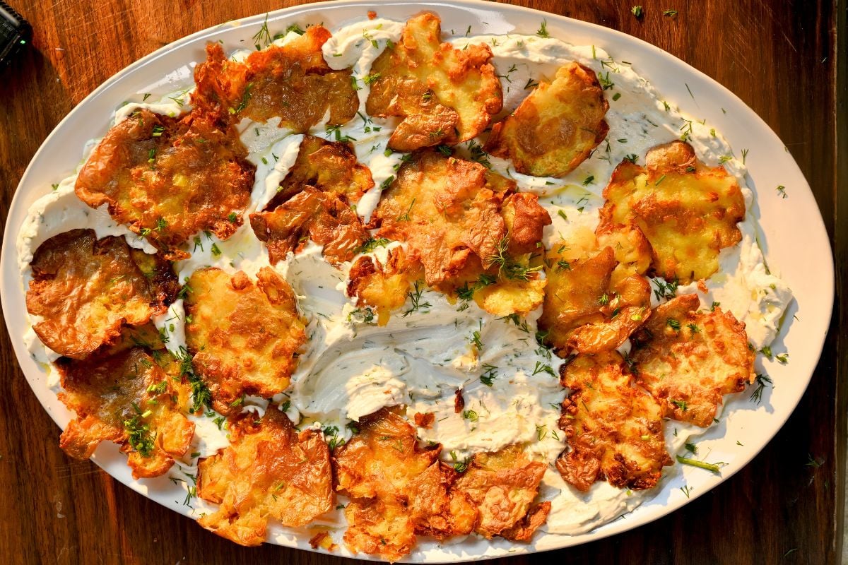 A top-down view of a large oval platter featuring smashed, deep golden-brown crispy potatoes arranged on a generous bed of creamy dill yogurt sauce, garnished with more fresh chopped dill.