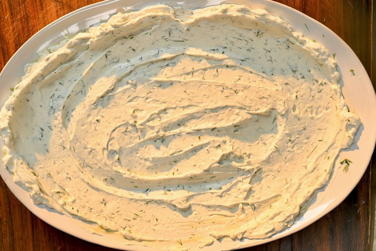 A top-down view of a creamy white yogurt sauce mixed with fresh chopped dill, spread smoothly in swirls across a large oval serving platter.