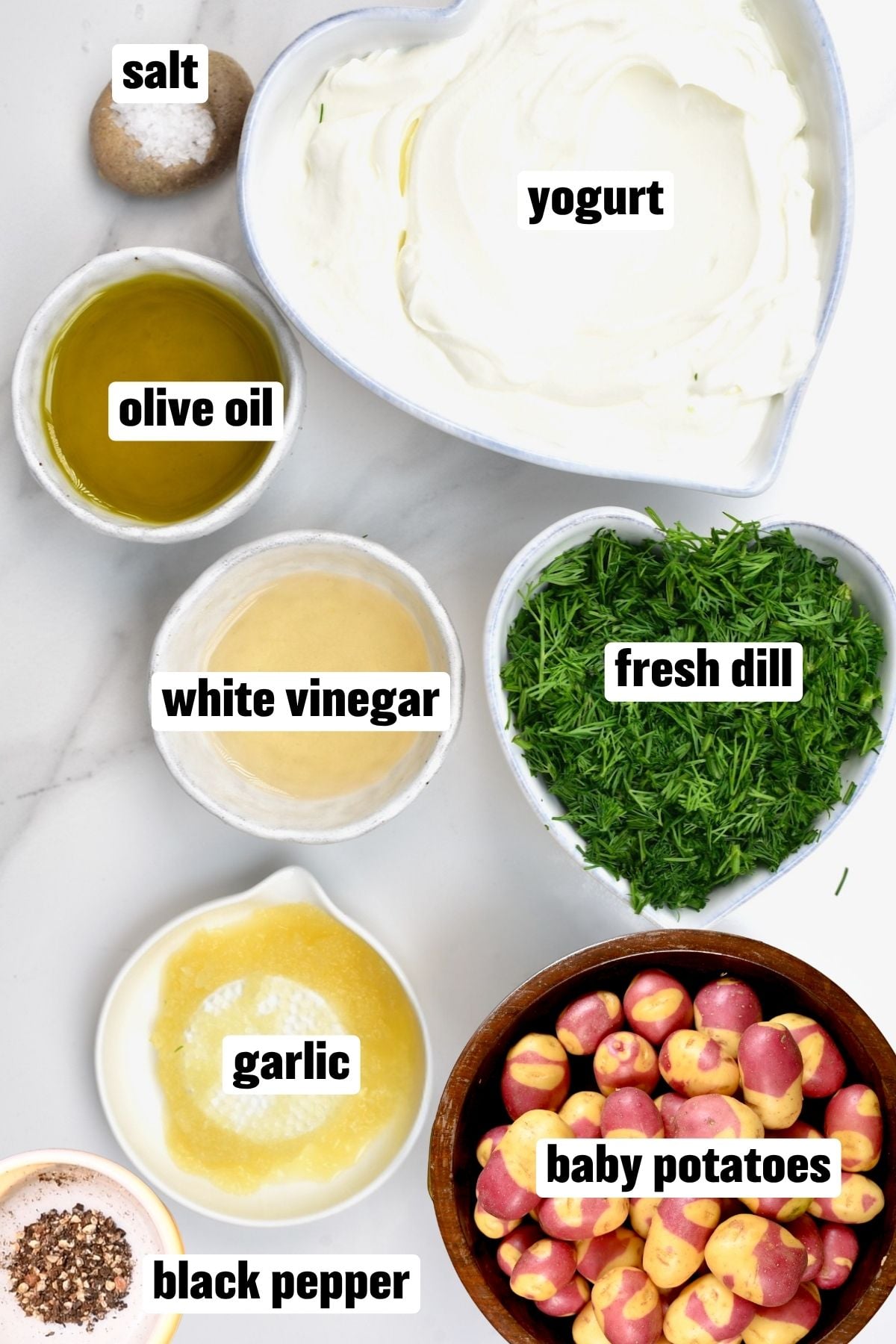 An ingredient flat lay arranged on a white surface featuring plain yogurt, fresh dill in a heart-shaped bowl, baby red potatoes in a wooden bowl, alongside small bowls containing olive oil, white vinegar, minced garlic, salt, and black pepper.