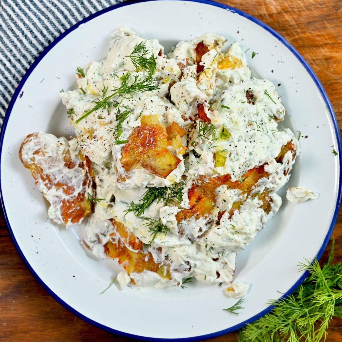 A close-up, top-down view of a serving of crispy smashed potatoes fully tossed and coated in creamy dill yogurt dressing, served on a white enamel plate with a blue rim and garnished with fresh dill sprigs and cracked black pepper.