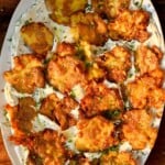 An overhead, close-up shot of a large oval platter filled with golden, extra-crispy smashed potatoes layered over a smooth dill yogurt sauce and garnished with fresh chopped herbs.