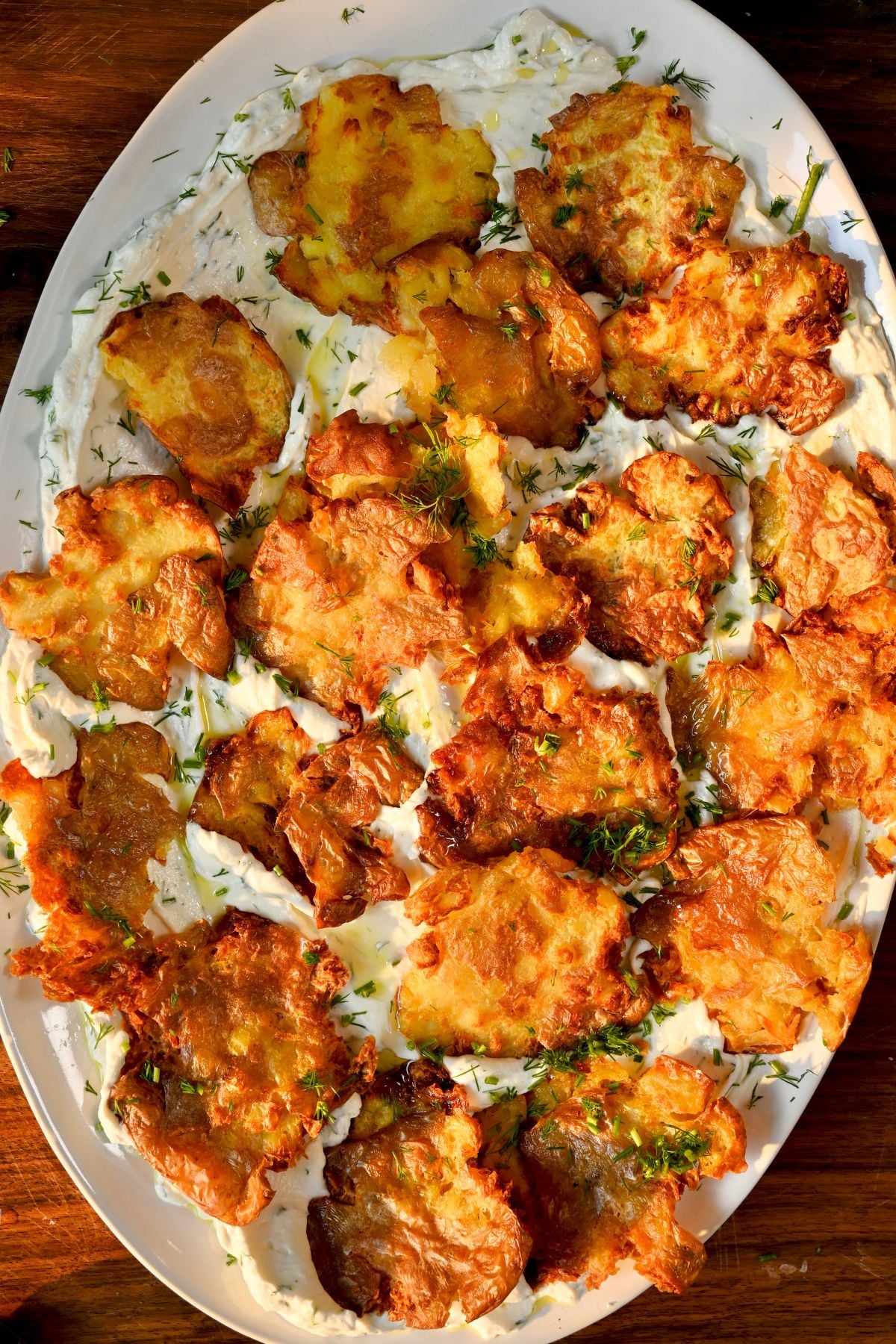 A full vertical view of an oval serving platter filled with crispy, golden smashed potatoes arranged over a bed of creamy dill yogurt sauce and sprinkled with fresh chopped herbs.