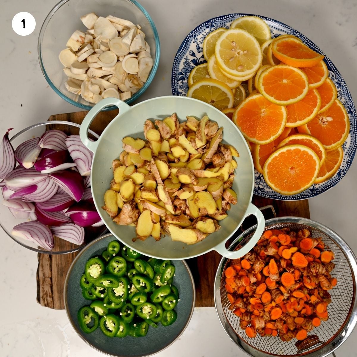 A top-down view of prepped fire cider produce organized in bowls, featuring sliced ginger, turmeric, horseradish root, red onion wedges, sliced jalapeños, lemon wheels, and orange wheels.