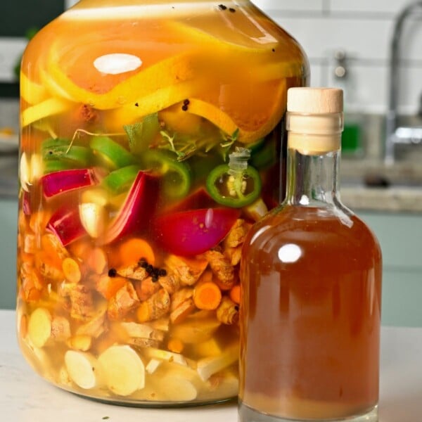 A close-up view of a cork-stoppered bottle of finished fire cider alongside a large glass jar filled with infusing roots, jalapeños, onions, garlic, and citrus slices.