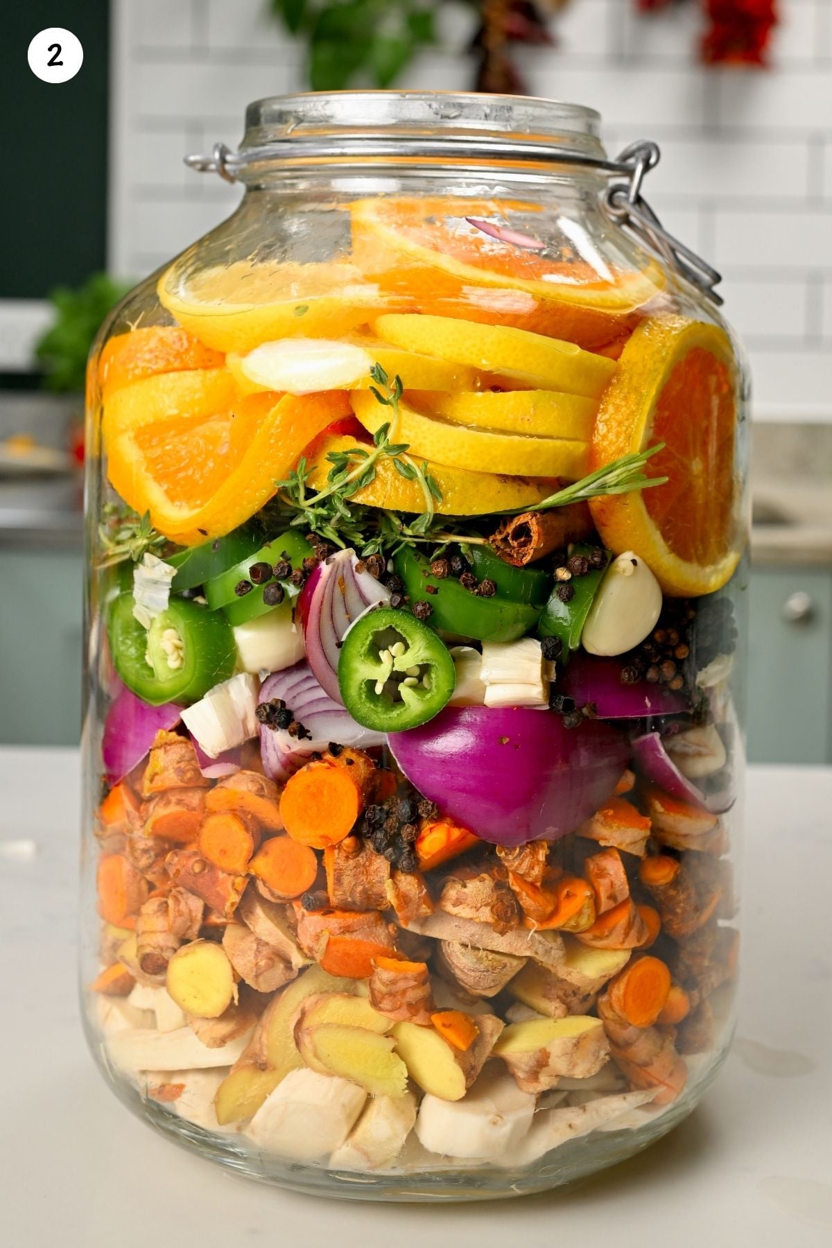 A large glass jar neatly packed with layers of roots, onions, garlic, jalapeños, whole spices, herbs, and citrus slices.