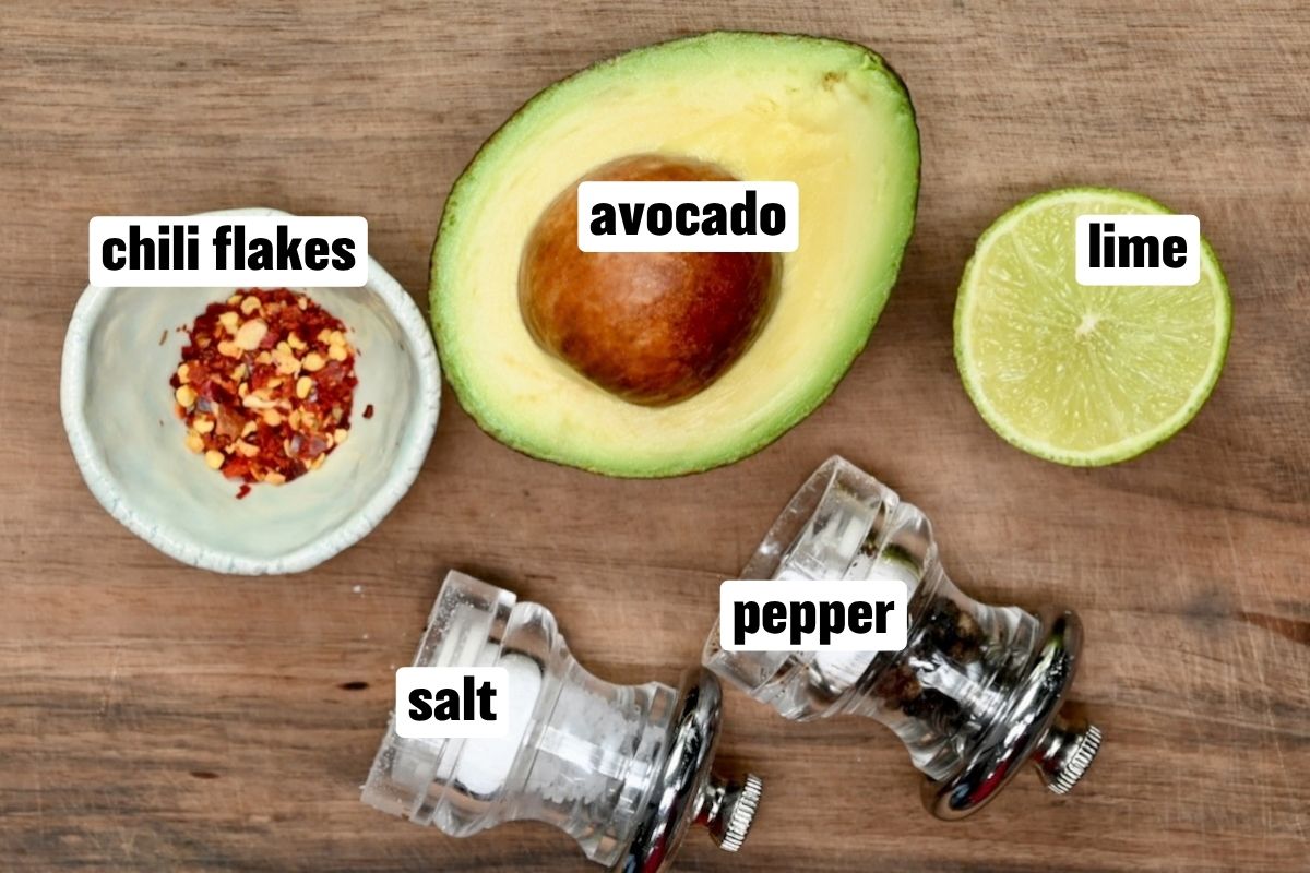 An overhead ingredient flat lay arranged on a wooden surface featuring half an avocado with its pit intact, half a fresh lime, red chili flakes in a small ceramic bowl, and clear glass salt and pepper shakers lying on their sides.