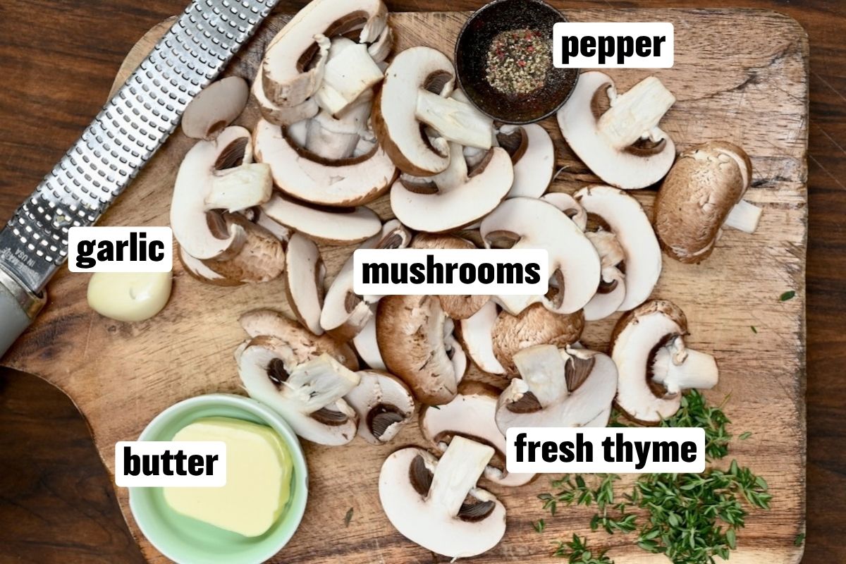 An overhead ingredient flat lay arranged on a rustic wooden board featuring sliced brown cremini mushrooms, butter in a small green dish, fresh sprigs of thyme, a clove of garlic next to a Microplane grater, and a small pinch bowl of black pepper.
