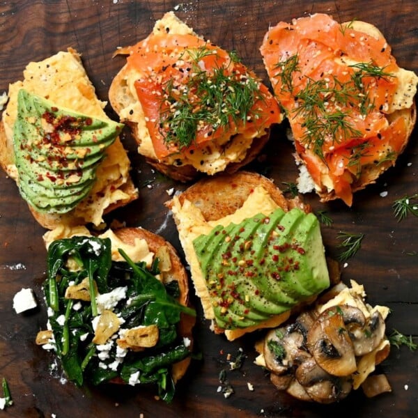 A top-down shot of multiple slices of toasted bread topped with fluffy scrambled eggs and various toppings, including fanned avocado with red chili flakes, smoked salmon with fresh dill and seeds, sautéed spinach with feta and crispy garlic, and sautéed mushrooms, arranged on a dark wooden board.