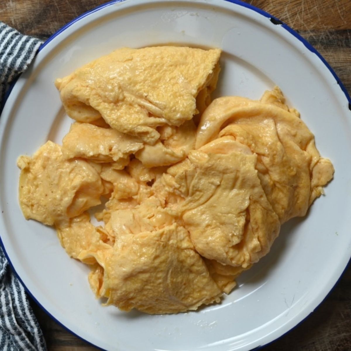 A top-down view of soft, fluffy scrambled egg curds served on a white enamel plate with a blue rim resting on a wooden table next to a striped cloth napkin.
