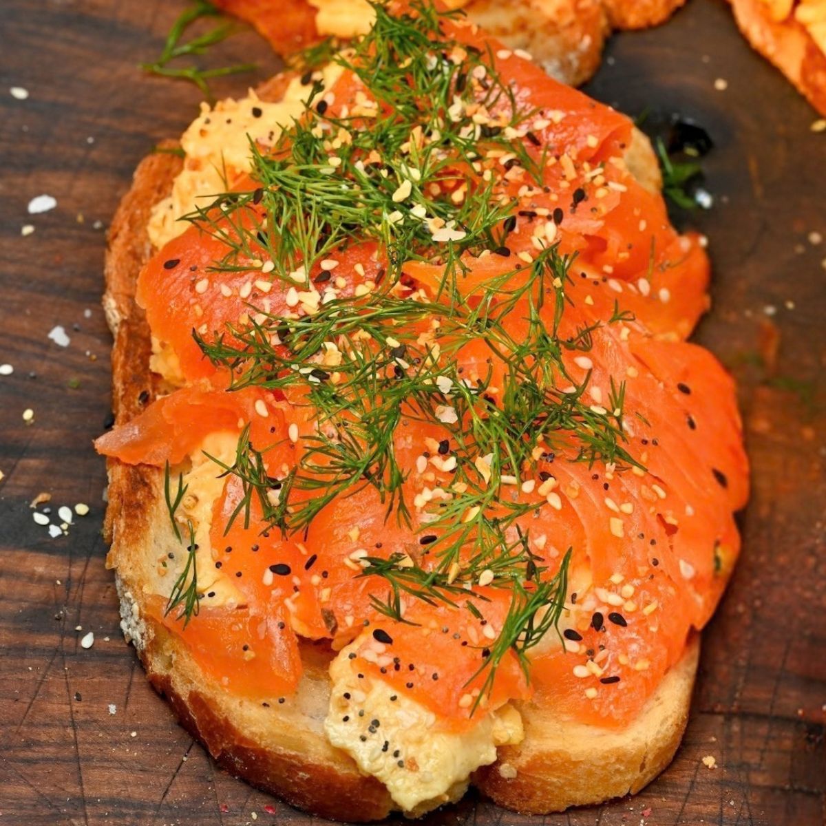 A top-down shot of a slice of toasted sourdough bread layered with soft scrambled eggs and thinly sliced smoked salmon, garnished with fresh dill fronds and everything bagel seasoning on a wooden board