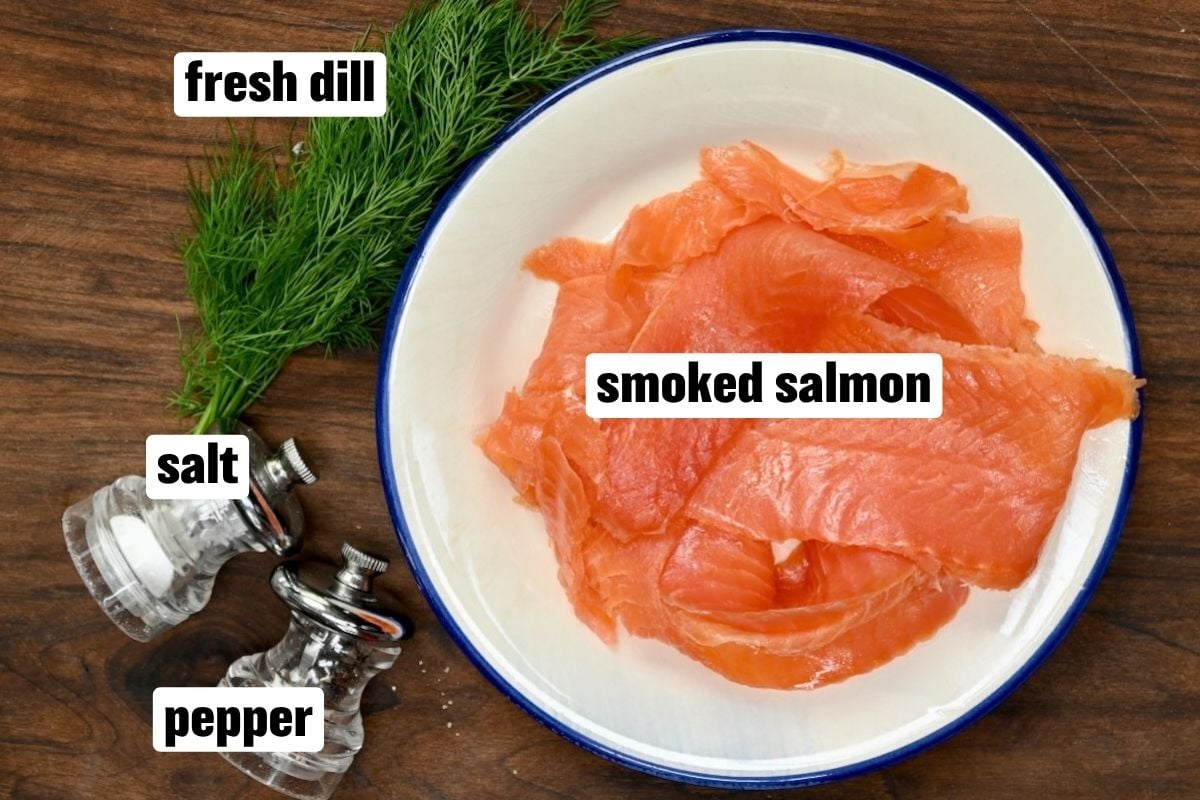 An overhead ingredient flat lay arranged on a wooden background featuring thinly sliced smoked salmon on a round white plate with a blue rim, fresh dill sprigs, and small clear glass salt and pepper shakers